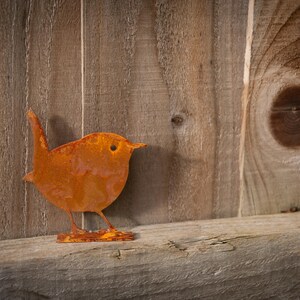 Close-up of wren fence topper with authentic rust finish, set against rustic fence to show its fine profile and textured patina. This minimalist metal bird sculpture adds instant woodland charm to courtyards, balconies and allotments. Built to weather gracefully all year round, the wren brings depth, movement and natural character to modern, farmhouse and cottagecore garden styles.