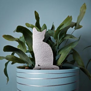 May include: A silver metal cat silhouette sitting in a blue planter with green leafy plants.