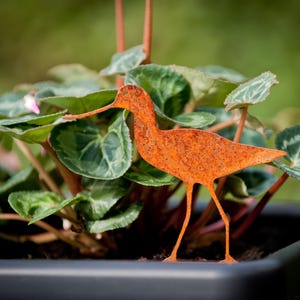 May include: A rusty orange metal bird sculpture in the shape of a wading bird, standing in a pot of green plants.