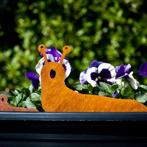 May include: A metal garden ornament depicting a large orange slug and two smaller slugs. The slug art is positioned in front of purple and white pansies in a black planter. The background is a blurred green bush.