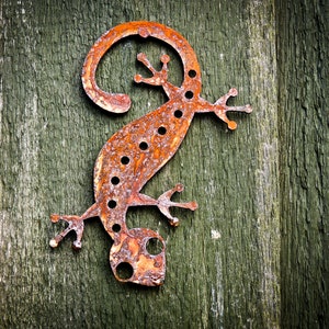 May include: A decorative, rusty metal lizard sculpture with a curled tail and small feet. The lizard features several circular cutouts. It is mounted on a weathered, green wooden surface.