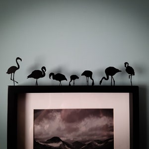 May include: A black metal silhouette art piece featuring seven flamingos in various poses, positioned along the top edge of a black picture frame that contains a black and white landscape photograph of mountains under a cloudy sky.