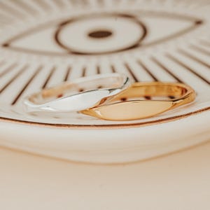 May include: Two simple, minimalist rings, one silver and one gold, are stacked on a white plate with a black and white eye design.