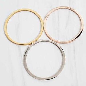 May include: Three metal bangles in gold, rose gold, and silver are arranged on a white marble surface. The bangles are circular and have a smooth, polished finish. The gold bangle is at the top left, the rose gold at the top right, and the silver at the bottom.