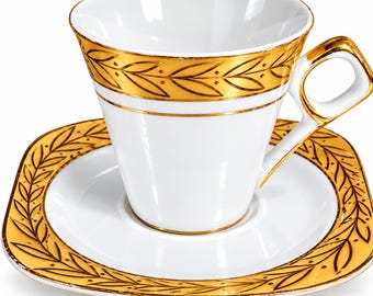 May include: A white porcelain teacup and matching square saucer featuring a gold-colored laurel leaf pattern along the rim and handle. The cup has a flared top edge and a geometric handle, while the saucer has rounded corners.