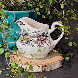 May include: A white ceramic creamer with a floral pattern featuring red, pink, yellow, and blue flowers. The creamer has a curved spout and a handle. It sits on a round wooden slice, with a turquoise vase in the background.
