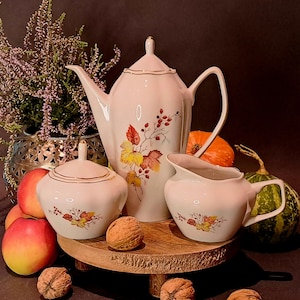 Puede incluir: Juego de té de porcelana blanca con tetera, azucarero y jarra de crema. Cada pieza está decorada con un borde dorado y un diseño floral otoñal. Se muestra con manzanas, nueces y calabazas.