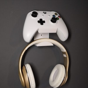 May include: White video game controller and gold headphones mounted on a white wall mount.