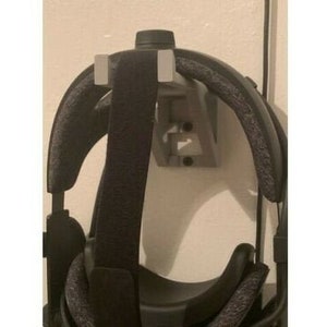 May include: A black VR headset with a grey wall mount. The headset has a padded headband and a strap that goes over the head.