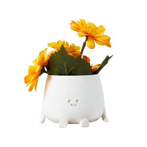 May include: A white ceramic planter shaped like a smiling face with small legs and arms, holding a bouquet of yellow flowers.
