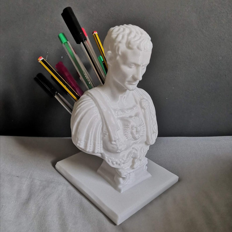 May include: A white bust of a Roman emperor, used as a pen holder. The bust is on a square base and holds several pens and pencils. The pens and pencils are various colors, including black, green, pink, and yellow.
