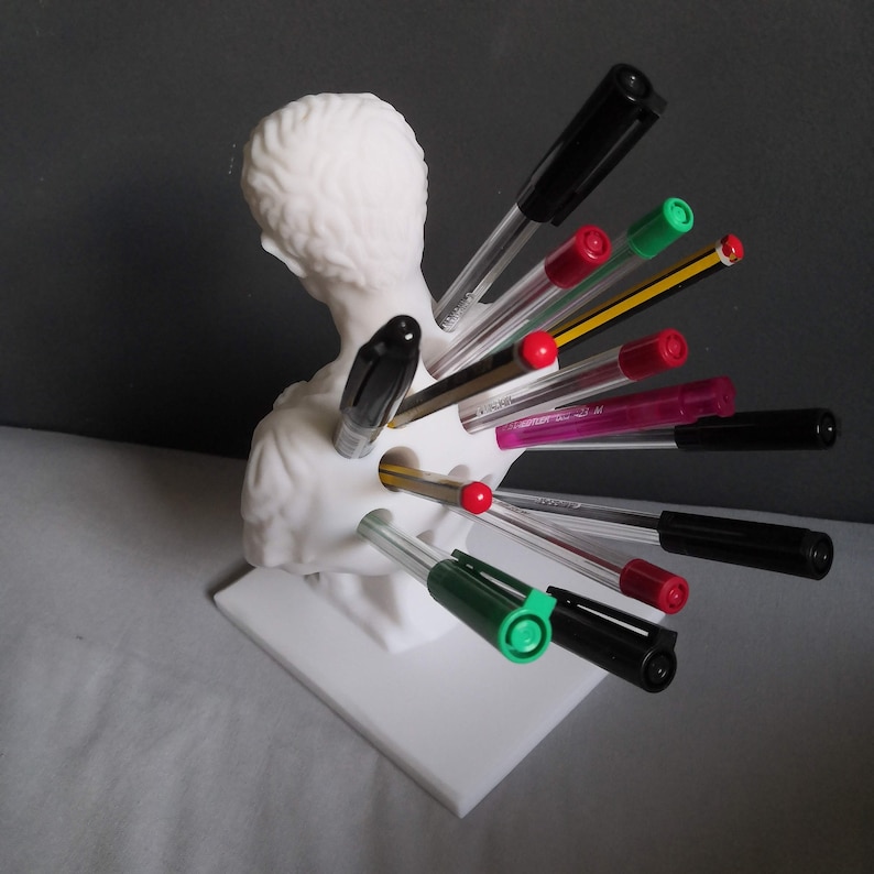 May include: A white bust-shaped pen holder with multiple pens and pencils inserted. The pens are various colors, including black, red, green, and purple. The holder is on a white square base.
