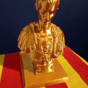May include: A gold-colored bust of a man, possibly a historical figure, with detailed facial features and attire. The bust is mounted on a square base. The background is a solid blue.