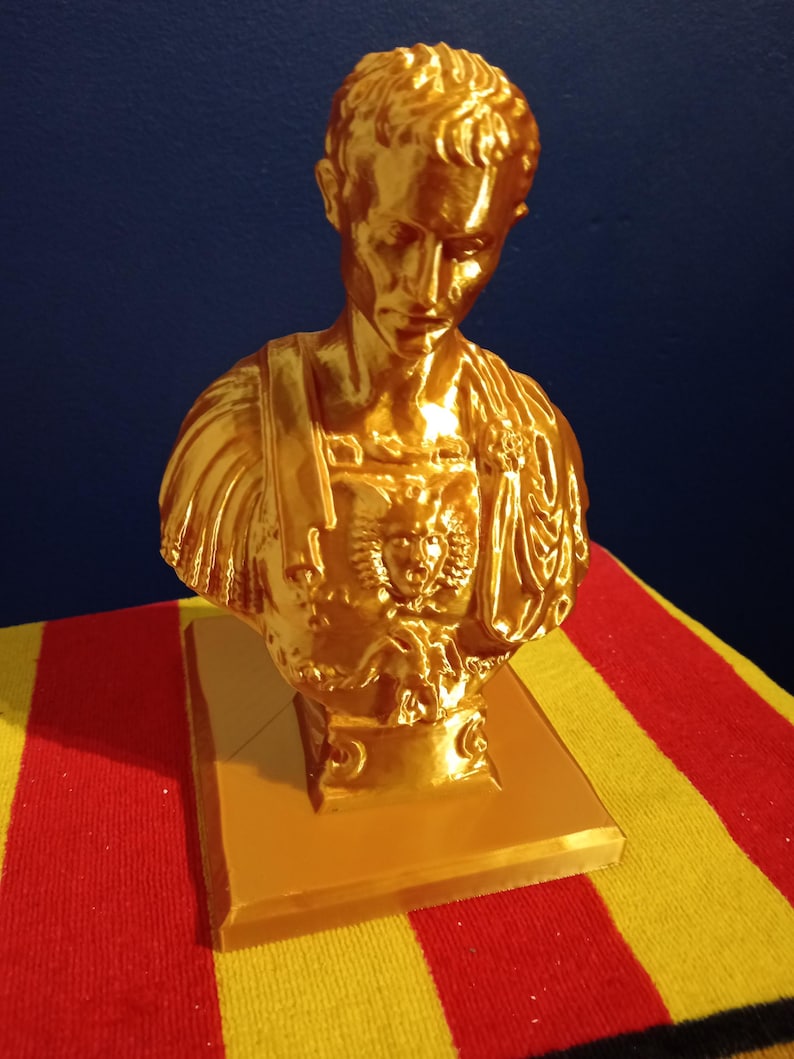 May include: A gold-colored bust of a man, possibly a historical figure, with detailed facial features and attire. The bust is mounted on a square base. The background is a solid blue.