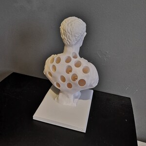 May include: A white bust sculpture with a unique design. The sculpture features a classical figure with numerous circular cutouts in the chest and back. It is mounted on a rectangular base, showcasing a modern artistic interpretation of a classical form.