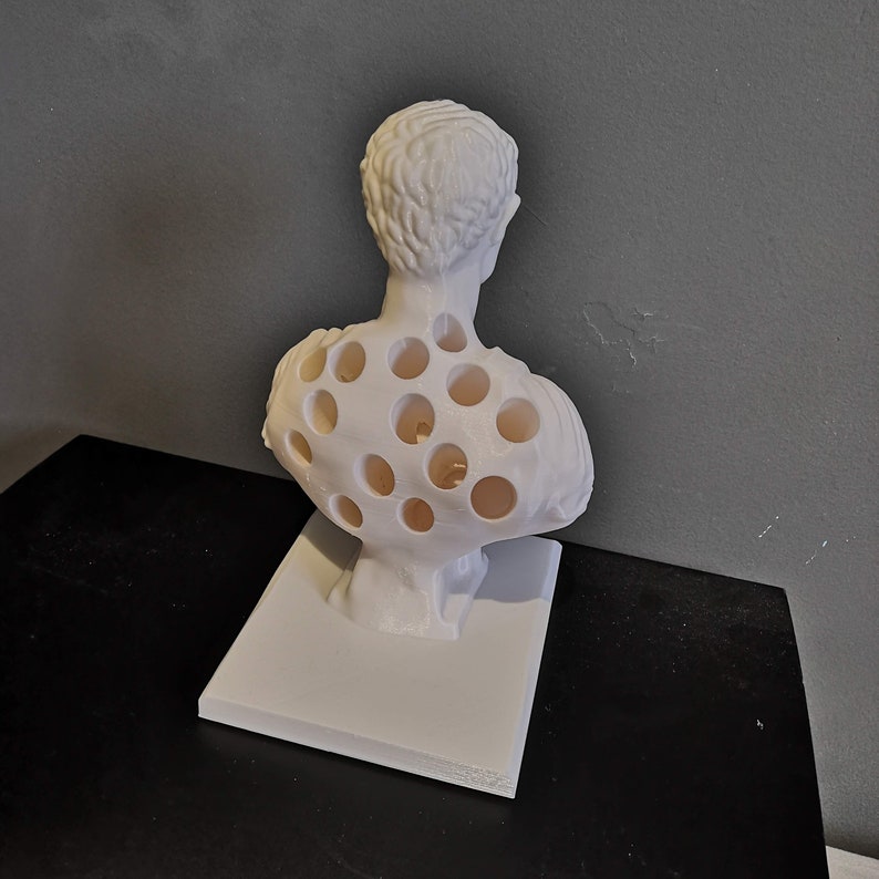 May include: A white bust sculpture with a unique design. The sculpture features a classical figure with numerous circular cutouts in the chest and back. It is mounted on a rectangular base, showcasing a modern artistic interpretation of a classical form.