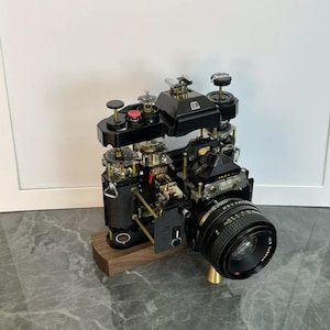 May include: A disassembled black 35mm film camera mounted on a dark wood base. The internal mechanical components, including gears, springs, and a red shutter button, are exposed. A black lens is attached to the front of the camera body.