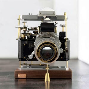 Könnte beinhalten: Eine zerlegte Vintage-Kamera auf einem dunklen Holzsockel. Die Kamera zeigt freiliegende schwarze und silberne mechanische Komponenten, Messingstützstangen und ein zentrales Objektiv. Eine kleine rechteckige Messingplatte ist am Sockel befestigt.