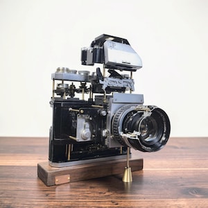 Könnte beinhalten: Eine zerlegte Vintage-Petri-Kamera, die auf einem rechteckigen Holzsockel montiert ist. Die internen mechanischen Komponenten, einschließlich Zahnrädern, Hebeln und der Objektiveinheit, sind freigelegt und vertikal getrennt. Eine kleine Kupferplatte befindet sich auf dem Sockel.