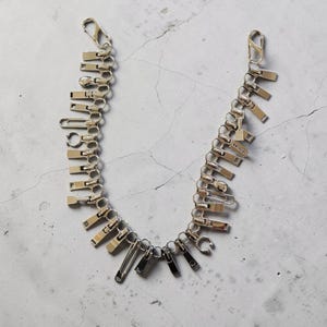 Puede incluir: Una pulsera de cadena plateada construida a partir de una serie de tiradores de cremallera metálicos interconectados, pequeños imperdibles y anillas circulares. El accesorio cuenta con un cierre de mosquetón en cada extremo en forma de U.