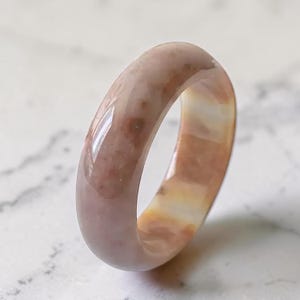 Op de afbeelding: Een gladde, gepolijste bandring gemaakt van gevlekt steenmateriaal. De ring heeft een afgerond profiel met een glanzende afwerking en toont een mix van zachte beige, gedempte roze en lichtbruine tinten met natuurlijke variaties in het oppervlak.