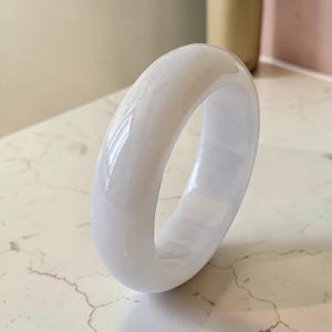 Puede incluir: Un brazalete rígido de color blanco, liso y sólido, con un acabado pulido y brillante. El accesorio circular presenta un perfil grueso y redondeado con una apariencia translúcida, reflejando la luz sobre su superficie curva para resaltar su color blanco uniforme.