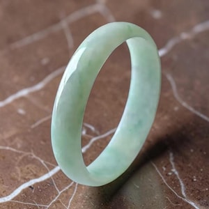 Op de afbeelding: Een gladde, gepolijste armband van lichtgroene jade. De cirkelvormige band heeft een doorschijnend uiterlijk met subtiele variaties in kleurintensiteit en een glanzende afwerking die het licht reflecteert over het afgeronde oppervlak.