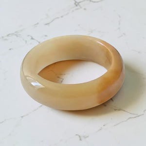 Pode incluir: Uma pulseira tipo bracelete lisa e polida, feita de uma pedra translúcida de cor de mel. A peça de joalharia circular apresenta um perfil arredondado com variações subtis de tom, variando do creme pálido a um tom âmbar dourado quente.