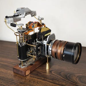 Könnte beinhalten: Eine zerlegte Canon AE-1 Kamera auf einem rechteckigen Holzsockel. Das silberne Gehäuseoberteil schwebt über dem schwarzen Innenchassis und gibt den Blick auf filigrane Messingstangen, Kupferdrähte und ein markantes schwarz-kupfernes Objektiv frei.