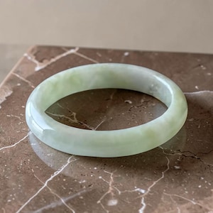 May include: A smooth, polished bangle bracelet crafted from pale green jade. The circular accessory features a translucent quality with subtle variations in color, showcasing a rounded profile and a continuous, seamless band design.