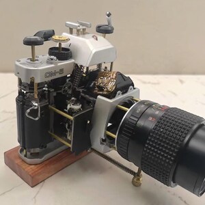 Könnte beinhalten: Ein zerlegtes Kameramodell mit der Aufschrift CM-5, das freiliegende interne mechanische Komponenten, Zahnräder und Schaltkreise zeigt. Das Gerät ist auf einem rechteckigen Holzsockel montiert und hat ein schwarzes, texturiertes Objektiv.