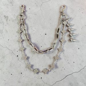 May include: A silver-toned multi-strand chain accessory featuring a thick rectangular link chain and a secondary chain composed of small, rhinestone-encrusted clover-shaped links. The piece includes two lobster clasps and a dangling spike charm.