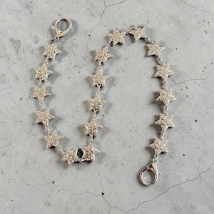 Könnte beinhalten: Ein silberfarbenes Kettenarmband mit einer Reihe verbundener sternförmiger Anhänger, die jeweils mit kleinen klaren Strasssteinen besetzt sind. Das Armband verfügt an beiden Enden über einen Karabinerverschluss zum Befestigen.