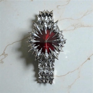 May include: A distinctive wristwatch with a bold design. The watch has a deep red face and a silver-coloured, spiky metal band and casing. The watch has a textured, almost brutalist aesthetic, with a modern and edgy appeal.