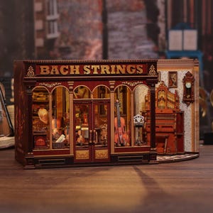 Può includere: Un modellino in legno di un negozio di musica intitolato BACH STRINGS, caratterizzato da una vetrina rosso scuro con scritte dorate, interni illuminati con violini, un grammofono, un organo e un orologio da parete sul lato.