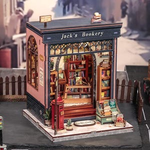 Könnte beinhalten: Ein Miniatur-Buchladen-Modell mit der Aufschrift Jack's Bookery, mit rosafarbener Fassade, dunkelblauen Verzierungen und einer offenen Front, die detaillierte Regale voller winziger Bücher, einen roten Briefkasten und eine Fußmatte zeigt.