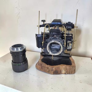 Könnte beinhalten: Ein zerlegtes, schwarzes Minolta X-300s Kameragehäuse, montiert auf einem Naturholzsockel mit Messingstützstangen, begleitet von einem separaten schwarzen Kameraobjektiv. Die internen Komponenten sind freigelegt und zeigen den Metall-Objektivanschluss.