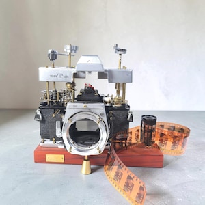 Könnte beinhalten: Eine demontierte Minolta SRT 101 Kamera mit freiliegenden internen Zahnrädern und Mechanismen, montiert auf einem rechteckigen Holzsockel. Ein 35mm-Film mit sichtbaren Bildern ist neben dem Kameragehäuse abgerollt, mit silbernen und schwarzen Komponenten.