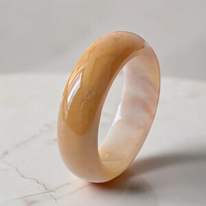 Puede incluir: Un anillo liso y pulido con perfil redondeado, hecho de un material translúcido de color melocotón cálido con un sutil veteado blanco nublado en toda su estructura, creando una apariencia suave y brillante con reflejos de luz en la superficie.