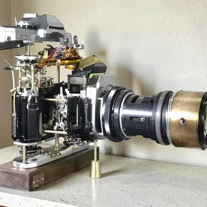 Puede incluir: Una cámara Canon AE-1 desmontada montada sobre una base rectangular de madera. Los componentes mecánicos internos, engranajes y cableado están expuestos. Un objetivo negro grande con un anillo frontal dorado está unido al cuerpo de la cámara.