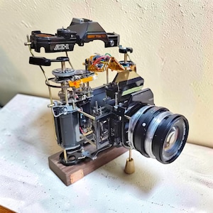 Puede incluir: Una cámara Canon AE-1 desmontada montada sobre una base rectangular de madera. Los componentes mecánicos internos, engranajes y cableado están expuestos y suspendidos en una vista explosionada. El cuerpo negro, el objetivo y la carcasa son visibles.