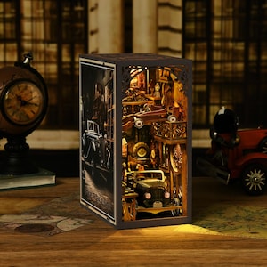 Könnte beinhalten: Ein beleuchtetes Holz-Diorama, das eine Oldtimer-Werkstatt und eine Straßenszene zeigt. Das detaillierte, lasergeschnittene Design zeigt ein klassisches Auto, Werkzeuge und architektonische Elemente und erzeugt ein warmes Leuchten. Das Diorama ist etwa 15 cm hoch.
