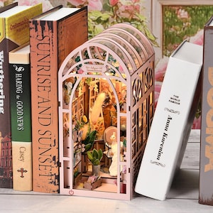 Könnte beinhalten: Ein Miniaturmodell eines rosa Gewächshauses mit Innenbeleuchtung, das eine detaillierte Innenszene mit Miniaturpflanzen und Figuren zeigt. Das Modell steht neben mehreren Büchern, darunter "Sunrise and Sunset" und "Anna Karenina".