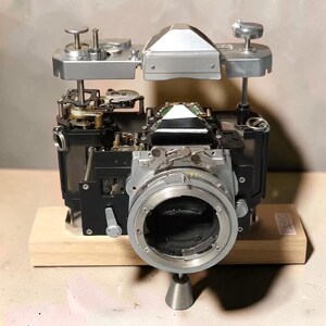 Puede incluir: Una cámara de película de 35 mm desmontada con sus componentes mecánicos internos expuestos. La placa superior plateada está elevada sobre el cuerpo negro de la cámara, revelando engranajes, palancas y la montura del objetivo sobre una base de madera.