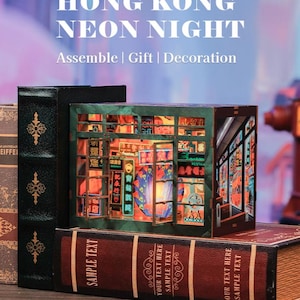 Könnte beinhalten: Ein Miniatur-Diorama-Bausatz mit dem Titel HONG KONG NEON NIGHT, Assemble | Gift | Decoration, bestehend aus einem dunkelgrünen Rahmen mit offenen Fenstern, die beleuchtete Neonschilder mit chinesischen Schriftzeichen zeigen, zwischen zwei buchförmigen Boxen.