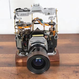 Könnte beinhalten: Eine zerlegte Canon AE-1 Kamera auf einem Holzsockel, die ihre internen mechanischen Komponenten, Zahnräder und Schaltkreise freilegt. Die silberne Deckplatte ist über dem Hauptgehäuse angehoben und zeigt die komplexe Technik des Geräts.