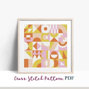 May include: A cross stitch pattern featuring a geometric design with orange, yellow, pink, and white shapes. The pattern is framed in a light wood frame.