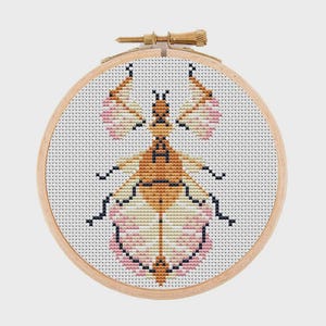 May include: A cross-stitch embroidery piece featuring a stylised insect with a tan body and pink-tinted wings, displayed in a circular wooden hoop. The design is stitched onto white Aida fabric with a visible grid pattern and a brass tension screw.