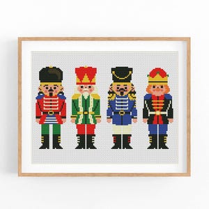 Op de afbeelding: Een kruissteekpatroon met vier notenkrakerfiguren op een rij, elk met een eigen uniform in rood, groen, blauw en zwart met gouden accenten, geplaatst tegen een witte rasterachtergrond en ingelijst in een lichthouten lijst.