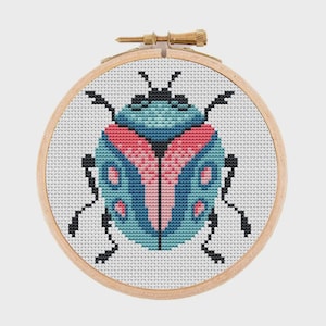 May include: A circular wooden embroidery hoop containing a cross-stitch pattern of a beetle. The beetle features a blue and teal body with pink wing accents and black legs, antennae, and head details, all stitched onto a white fabric background.